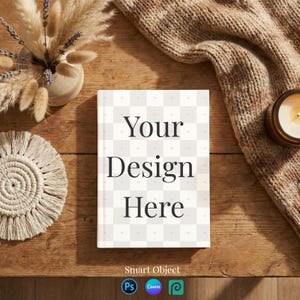 May include: A hardcover book with the text "Your Design Here" on a wooden surface. The book is surrounded by a candle, a knitted blanket, a vase with dried plants, and a macrame coaster. The image has a warm, inviting aesthetic.