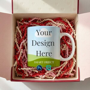 Op de afbeelding: Een witte keramische mok met de tekst "Your Design Here" staat in een geschenkdoos gevuld met rode en crèmekleurige papiersnippers. De mok heeft een blauwe luchtontwerp met een groen landschap aan de onderkant. De doos is roze met hartvormige uitsparingen.