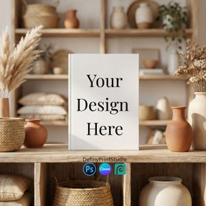 Puede incluir: Un libro blanco con el texto "Your Design Here" está sobre una estantería de madera. La estantería forma parte de una exhibición con jarrones decorativos, cestas y plantas secas. El libro es una maqueta.