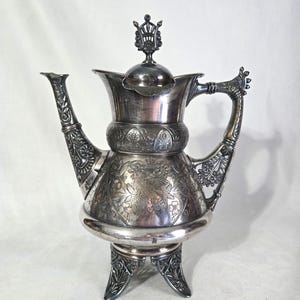 May include: Ornate silver teapot with intricate floral and scrollwork designs. The teapot features a decorative handle, spout, and a crown-shaped finial on the lid. It stands on three decorative legs.