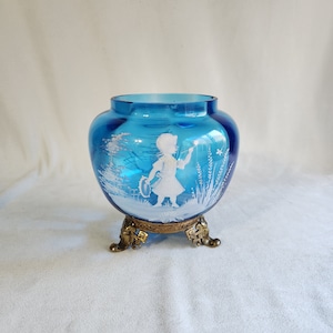 Peut inclure: Un vase en verre bleu avec une silhouette blanche d'une fille soufflant des bulles. Le vase a une base en métal doré avec trois pieds.