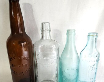 May include: Four vintage glass bottles, two clear and two light blue. The brown bottle on the left has the text "Heymann Br. & Co. Brewing Co. Inc. New York" printed on it. The clear bottle in the center has the text "The Monarch Brewing Co. St. Louis, Mo. U.S.A." printed on it. The light blue bottle on the right has the text "T.H. Kelly & Co. St. Louis, Mo." printed on it.
