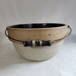 Op de afbeelding: Een witte keramische kom met een bruin geglazuurd interieur en een houten handvat. De kom heeft een draadrand en is ontworpen om voedsel te serveren of op te slaan.