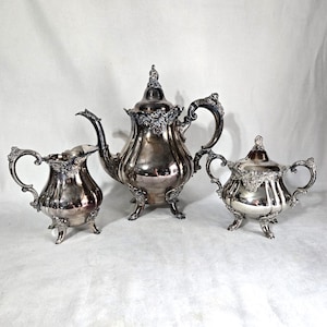 May include: A silver tea set including a teapot, creamer, and sugar bowl. The set has ornate detailing, curved handles, and sits on small feet. The teapot has a tall, decorative finial on the lid. The set is displayed on a white surface.