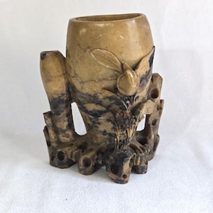 Puede incluir: Un jarrón de piedra tallada con un diseño naturalista. El jarrón es cilíndrico con una abertura ancha y está sostenido por una base que se asemeja a ramas de árboles. La piedra tiene un color beige y marrón moteado, con detalles tallados de hojas y una fruta.