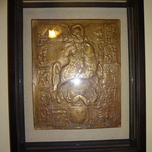May include: A framed bronze relief depicting a mounted figure, possibly a saint, with a sword and a shield. The relief is surrounded by a decorative border and is set against a beige background.