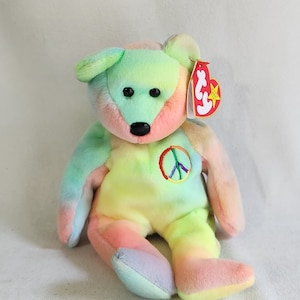 May include: A tie-dye rainbow colored beanie baby bear with a peace sign on its chest. The bear has black eyes and a black nose. The beanie baby tag is attached to the bear's right ear.