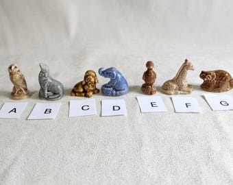 Może przedstawiać: Zestaw siedmiu małych ceramicznych figurek różnych zwierząt, w tym sowy, foki, psa, słonia, ptaka, żyrafy i lamparta. Figurki są ułożone w rzędzie na białym tle, z literami od A do G pod każdą figurką.