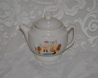 May include: White ceramic teapot with a lid, featuring a hand-painted scene of a fireplace with two chairs and a cat. The teapot has a round body and a small spout.