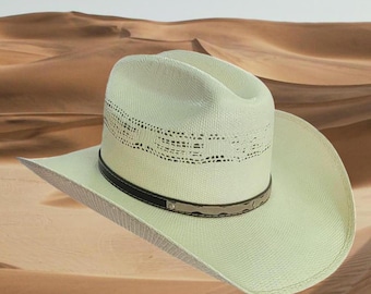 Puede incluir: Un sombrero vaquero de paja blanco con una banda negra y un diseño decorativo. El sombrero tiene un ala ancha y una corona alta.