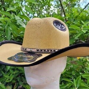 May include: A tan straw cowboy hat with a black band and a brown leather trim. The hat has a scorpion logo on the front.