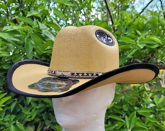 May include: A tan straw cowboy hat with a black band and a brown leather trim. The hat has a scorpion logo on the front.