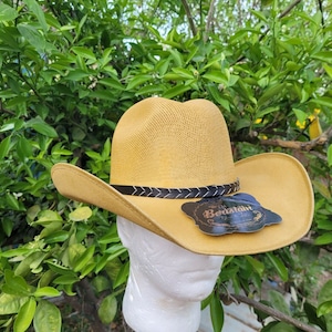 Op de afbeelding: Een gele strooien cowboyhoed met een zwarte gevlochten band en een bruin leren label met de tekst "The Old Bertsham Hat Co."