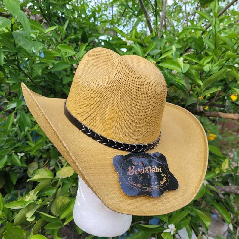 Op de afbeelding: Een beige strooien cowboyhoed met een zwarte leren band en een zwart label met de tekst "Via La Beristain".