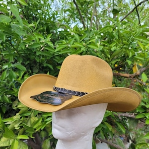 Op de afbeelding: Een beige cowboyhoed met een zwarte leren band en een klein zwart label met witte tekst. De hoed zit op een wit mannequinhoofd.