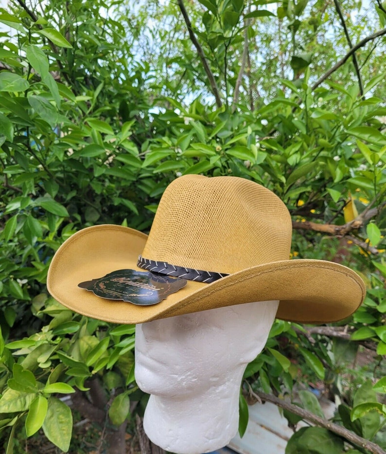 Op de afbeelding: Een beige cowboyhoed met een zwarte leren band en een klein zwart label met witte tekst. De hoed zit op een wit mannequinhoofd.