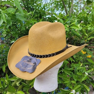 Op de afbeelding: Een beige cowboyhoed met een bruin leren band en een wit stikdetail. De hoed heeft een brede rand en een hoge kroon. De hoed heeft een label met de tekst "The  Benistaini".