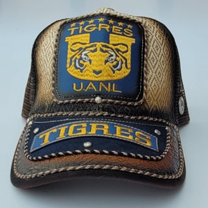 May include: A baseball cap with a mesh back and a leather bill. The front features a blue patch with the word "TIGRES" in yellow and a tiger logo. The cap has a brown and tan color scheme.
