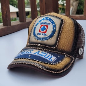 May include: A brown and tan baseball cap with a blue and white embroidered patch that says "DEPORTIVO CRUZ AZUL MEXICO". The cap also has a blue and white embroidered patch that says "CRUZ AZUL" on the front.