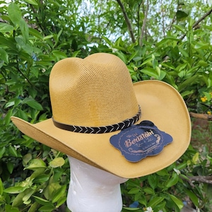 Op de afbeelding: Een gele strooien cowboyhoed met een zwarte leren band en een zwart label met de tekst "The Old Beretani" met een ontwerp van een veer en een ster.