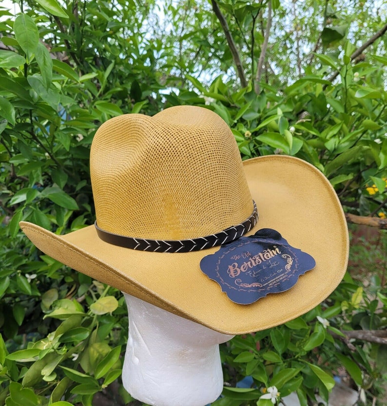 Op de afbeelding: Een gele strooien cowboyhoed met een zwarte leren band en een zwart label met de tekst "The Old Beretani" met een ontwerp van een veer en een ster.