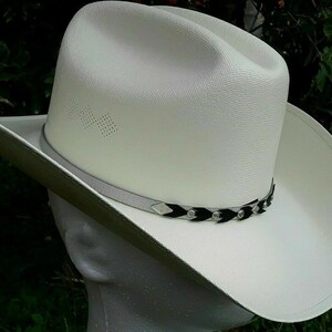 May include: A white cowboy hat with a black and silver braided band. The hat has a wide brim and a tall crown.