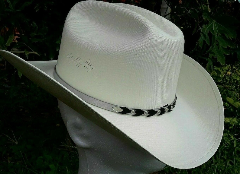 May include: A white cowboy hat with a black and silver braided band. The hat has a wide brim and a tall crown.