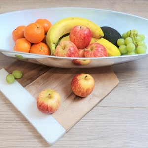 May include: A silver bowl filled with fruit, including bananas, apples, oranges, grapes, and an avocado. The bowl is sitting on a wooden chopping board with a white surface.