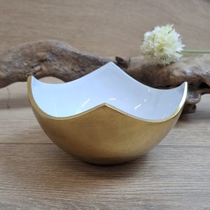 May include: A gold and white ceramic bowl with a unique, wavy design. The bowl is sitting on a wooden surface.