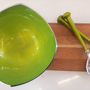 May include: A green ceramic bowl with a silver rim, a wooden serving board, and a green salad serving set with a fork and spoon.