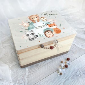 Può includere: Una scatola rettangolare in legno chiaro per ricordi. Il coperchio presenta un'illustrazione ad acquerello di un orso, una volpe, un procione, un gufo, un riccio e un coniglio. Il testo include il nome Mario, la data 20.11.2018 e puntini decorativi.