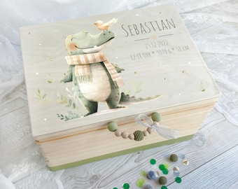 May include: A wooden keepsake box with a green and brown alligator design and a bird on top. The box has a wooden handle and a green and white ribbon. The box is personalized with the name "Sebastian" and the date "23.12.2022".