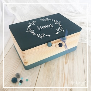 Könnte beinhalten: Eine dunkelblaue Holzkiste mit einem hellen Holzboden und einem türkisfarbenen Rand. Der Deckel ist mit einem weißen Blumenkranz und dem Namen "Henry" in weißen Buchstaben verziert. Die Kiste hat ein blaues Band und Holzperlen.