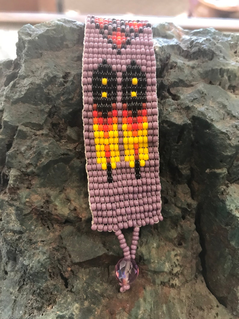 May include: A purple beaded bracelet with a colorful geometric design. The bracelet has a small purple crystal charm at the end.