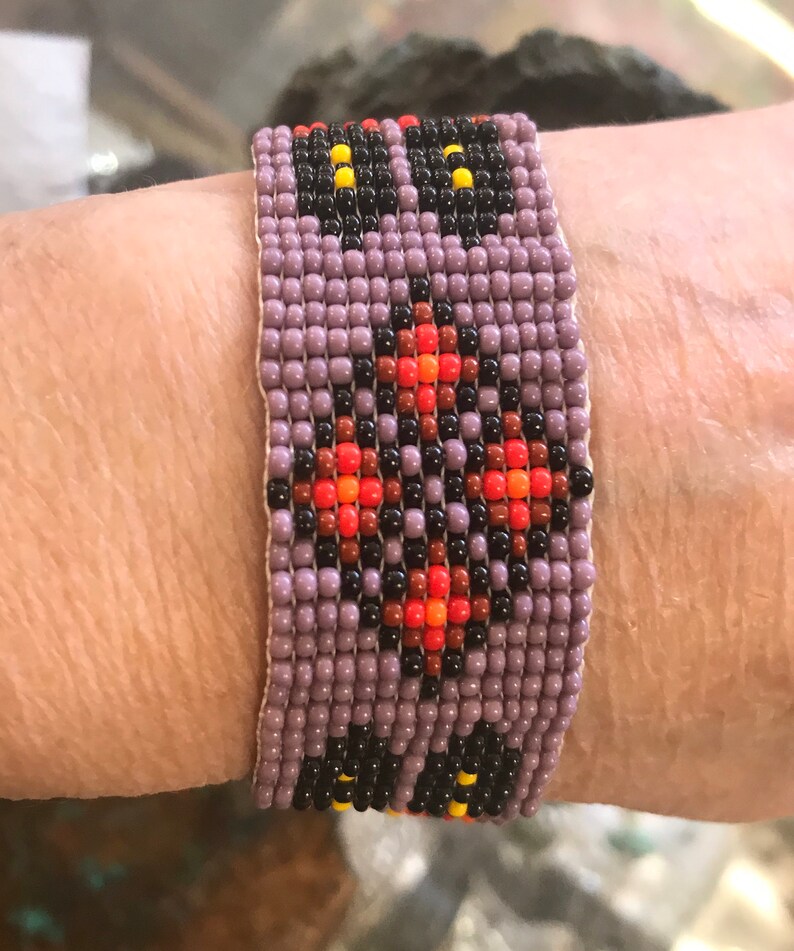May include: A beaded bracelet with a geometric pattern in shades of purple, black, orange, and red.