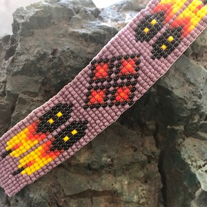 May include: A beaded bracelet with a purple base and a repeating pattern of black, orange, and yellow beads. The bracelet is made with small, round beads.