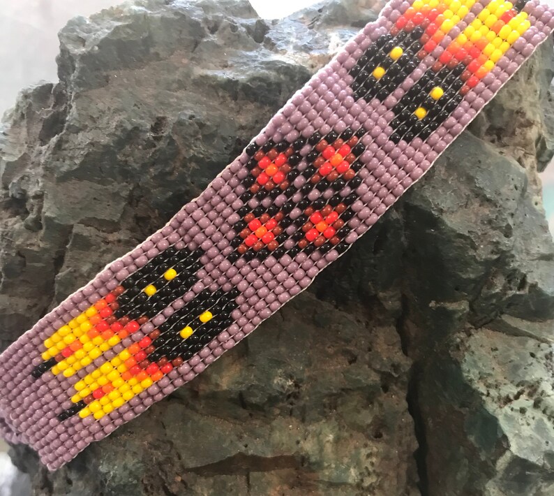 May include: A beaded bracelet with a purple base and a repeating pattern of black, orange, and yellow beads. The bracelet is made with small, round beads.