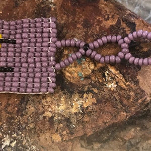 May include: A purple beaded bracelet with a colorful geometric pattern. The bracelet has a purple beaded closure.