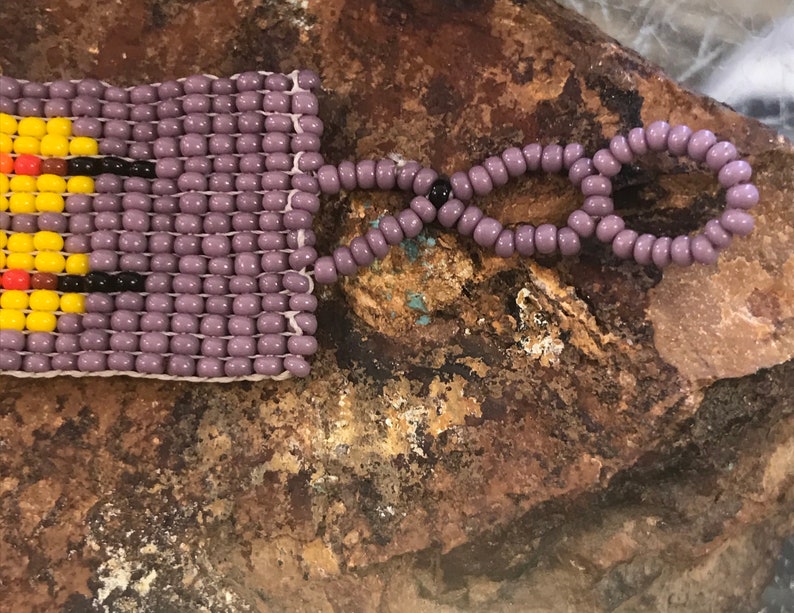 May include: A purple beaded bracelet with a colorful geometric pattern. The bracelet has a purple beaded closure.