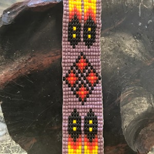 May include: A beaded bracelet with a purple background and a geometric design featuring black, yellow, orange, and red beads.