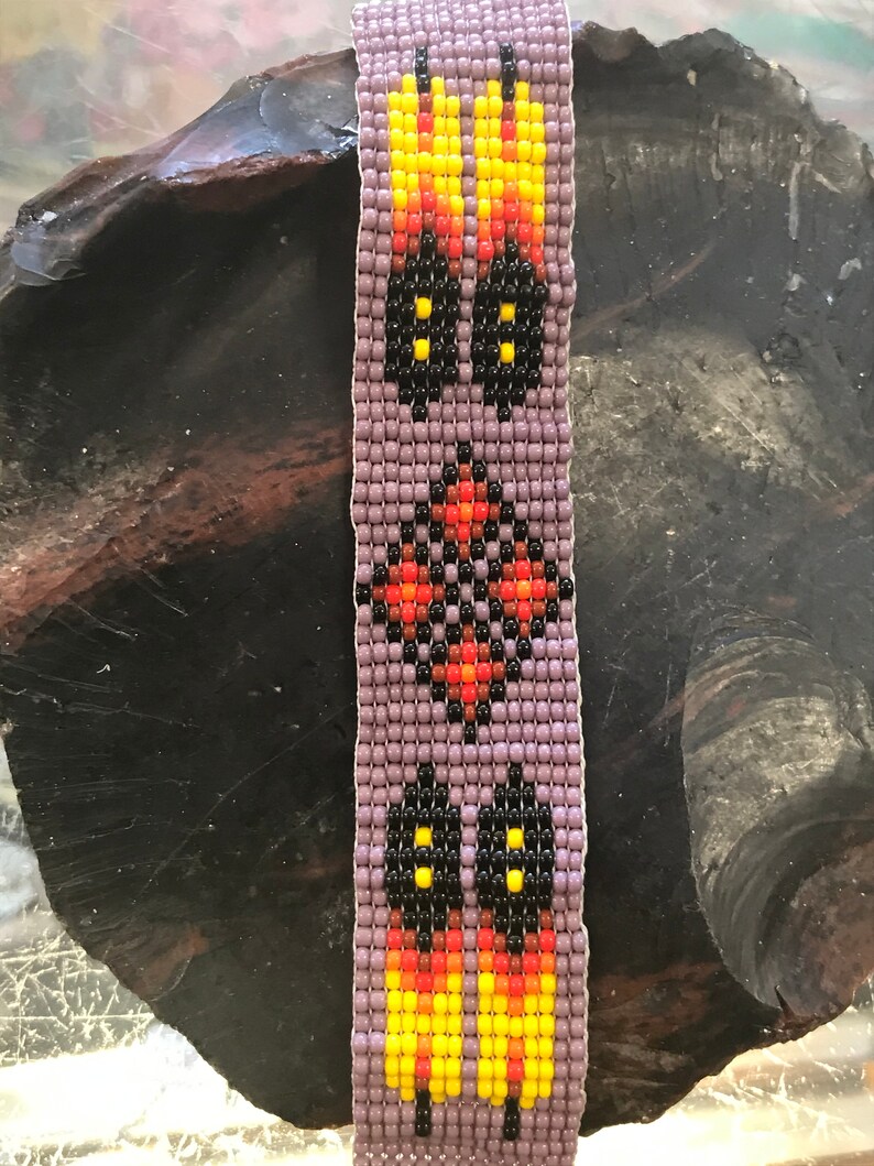 May include: A beaded bracelet with a purple background and a geometric design featuring black, yellow, orange, and red beads.