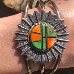 May include: Silver cuff bracelet with a central sunburst design. The center features a round inlay with orange and green geometric patterns. The bracelet has three curved bands and is likely made of sterling silver.