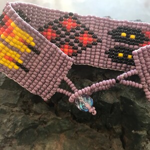 May include: A beaded bracelet with a purple base and colorful geometric patterns. The bracelet has a crystal charm.