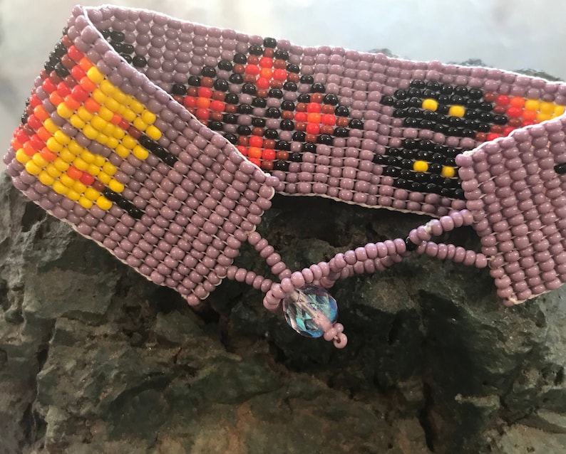 May include: A beaded bracelet with a purple base and colorful geometric patterns. The bracelet has a crystal charm.