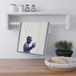 May include: A white kitchen towel with a blue border features a black and white image of a man wearing sunglasses and giving the middle finger.