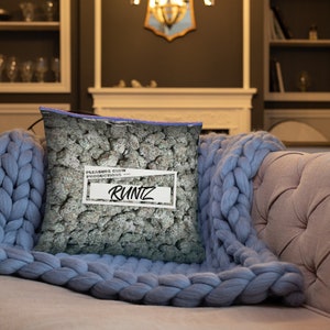 May include: A cushion with a graphic of a bag of cannabis buds. The cushion has a white label with the word "RUNTZ" in bold letters. The cushion is resting on a pink sofa and a blue knitted blanket.
