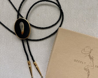 Op de afbeelding: Een bolo-das met een zwarte ovale hanger met een goudkleurige rand en een esdoornzaadje erin. De das bestaat uit een zwart gevlochten koord met goudkleurige metalen uiteinden. Ernaast staat een beige kartonnen doosje met de tekst DEEPLY SE.