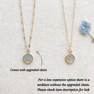 May include: Two gold necklaces with a small round pendant featuring a pressed blue flower. The necklace on the left has an upgraded chain. The necklace on the right has a standard chain. The text "Comes with upgraded chain" is below the left necklace. The text "For a less expensive option there is a necklace without the upgraded chain. Please check item description for link" is below the right necklace.