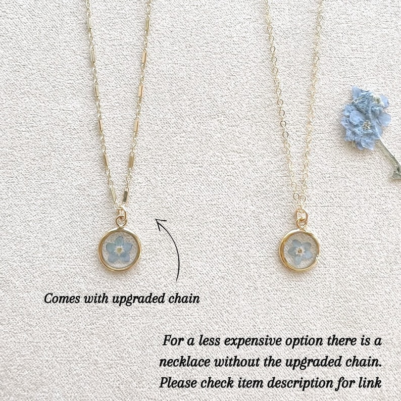 May include: Two gold necklaces with a small round pendant featuring a pressed blue flower. The necklace on the left has an upgraded chain. The necklace on the right has a standard chain. The text "Comes with upgraded chain" is below the left necklace. The text "For a less expensive option there is a necklace without the upgraded chain. Please check item description for link" is below the right necklace.