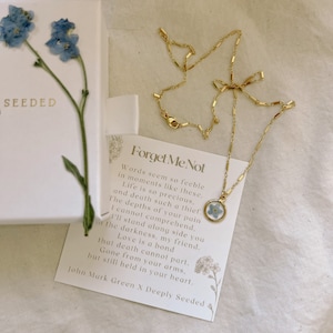 May include: Gold necklace featuring a forget-me-not flower pressed within a glass pendant. The necklace is displayed on a white card with the text "Forget Me Not" and a poem about love and loss.